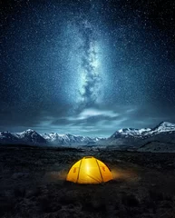 Wall murals Night blue Camping in the wilderness. A pitched tent under the glowing  night sky stars of the milky way with snowy mountains in the background. Nature landscape photo composite.