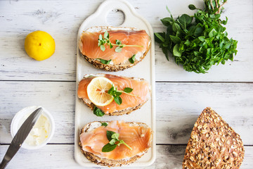 Sandwices with salted salmon and soft white bread cheese, decorated with a slice of lemon and fresh greens on an wooden background. Culinary background with appetizer of bread and red fish