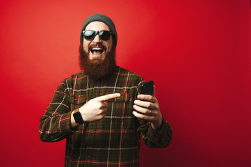 Cheerful amazed bearded hipster man pointing at phone over red background