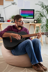 Young man in eyeglasses sitting on sofa and playing guitar