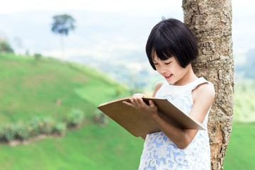 Fototapeta premium Cute little girl reading book on the mountain. back on tree, mountain behind, She has a look of enjoyment on her face and she looks very relaxed.