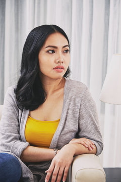 Attractive Filipino woman looking away and thinking while sitting on comfortable sofa in stylish living room
