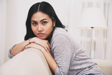 Side view of beautiful Filipino lady leaning on back of comfortable sofa and looking at camera while relaxing at cozy room at home