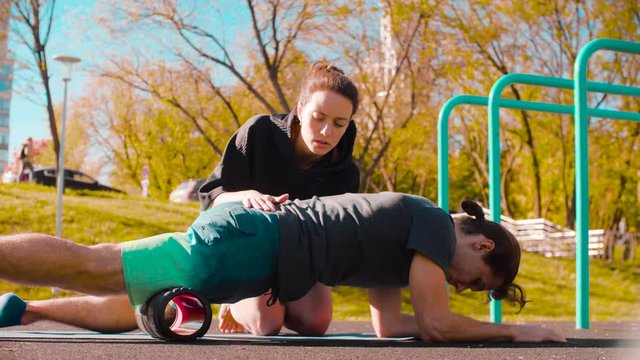 Man doing self massage of the legs with a roll. Woman teaching and correcting him. Sunny day, golden autumn