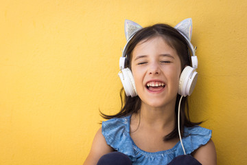Little girl singing with funny design headphones on. Cute kid listening to music over yellow wall background. Pop star wannabe, fun, joy, happy child concepts