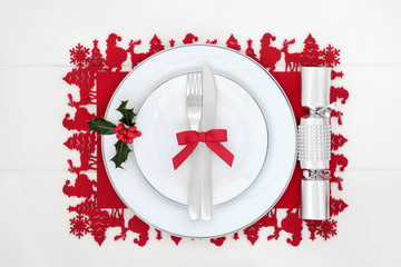 Christmas dinner table setting with porcelain plates, cutlery, diamond cracker, winter holly on red place mat on white wood table. Festive themed concept.