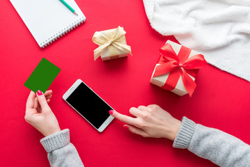 Female hands, woman holding a Business card, girl uses a white smart phone. Notepad on the red table, gifts boxes for the holidays, background with copy space for advertisement, top view