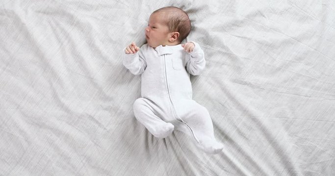 A newborn baby lying on the bed, looking into the camera and around on a white bed sheet, view from above. Infant knows the world, top view