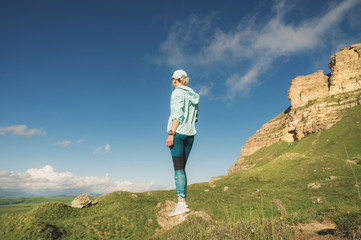 Attractive sports girl in a cap and headphones before jogging in a picturesque location next to the rocks at sunset. Workout outdoors. Back view