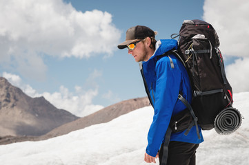 Fototapeta na wymiar Traveler in a cap and sunglasses with a backpack on his shoulders in the snowy mountains on the glacier against the sky and clouds. Traveler in a natural environment