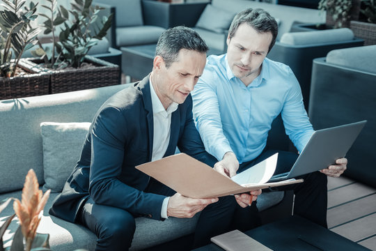 Give It Me. Serious Male Person Holding Laptop And Bowing Head While Sitting Near His Colleague