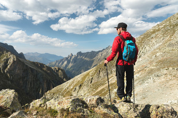 Fototapeta premium A bearded man in sunglasses and a cap with a backpack stands on top of a rock and looks into a rocky valley high in the mountains. The concept of tourism and easy trekking in the mountains outdoor
