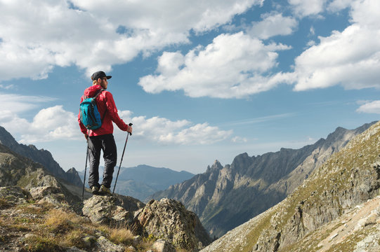 A bearded man in sunglasses and a cap with a backpack stands on top of a rock and looks into a rocky valley high in the mountains. The concept of tourism and easy trekking in the mountains outdoor