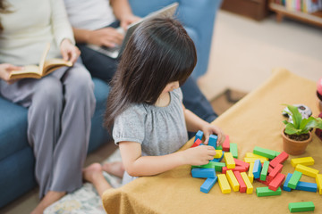 Family time Daughter laughing enjoying playing a game but her mother reading a book and her father working with laptop which sitting on sofa at home