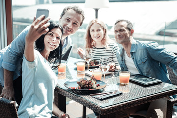 Say cheese. Charming female person raising her right arm while holding telephone and taking photo