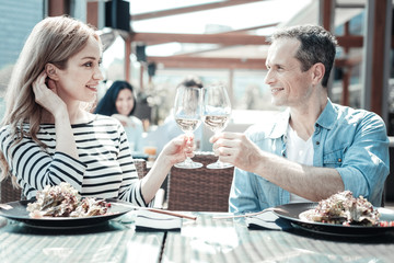 Sunny day. Pretty girl keeping smile on her face and touching hair while holding bocal with wine.