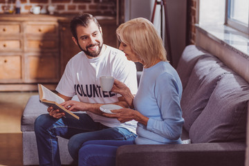 Relaxing together. Exuberant bearded volunteer reading a book for the aged woman and she drinking coffee