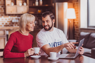 Modern technologies. Content dark-haired man smiling and teaching an old woman how to use the laptop