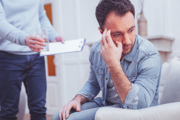 Life hardships. Portrait of emotionless bearded man looking away and touching his temples while sympathizing experienced psychiatrist holding a clipboard