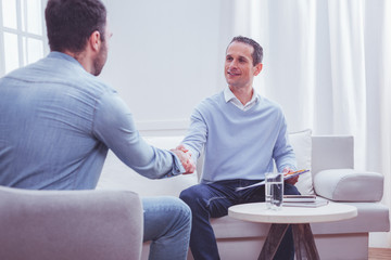 Glad to be helpful. Cheerful professional psychiatrist shaking hands with his satisfied client while sitting on the sofa and holding a clipboard in hand - obrazy, fototapety, plakaty