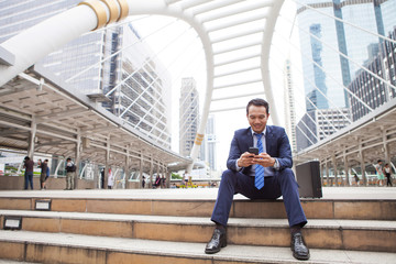 Businessman sitting on stair while looking on mobile phone, with office building background - business success, achievement, and win concepts