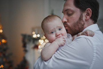 happy father kissing his baby daughter for the Christmas tree lights in the background. Family, fatherhood and holidays concept.