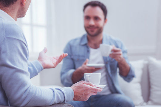 Pleasant meeting. Best positive friends holding cups and drinking tea while discussing latest events and sitting on sofa