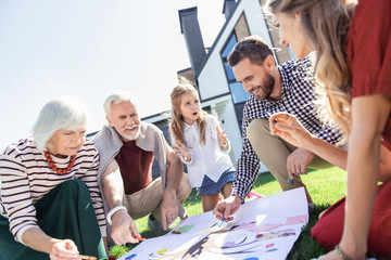 Give explanations. Pleased bearded man sitting near his wife and helping to draw picture