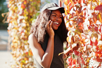 Stylish african american girl in cap posed at sunny autumn day against red leaves. Africa model woman.