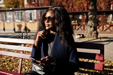Stylish african american girl in blue coat and sunglasses posed at sunny autumn day at bench and using her mobile phone. Africa model woman with smartphone.