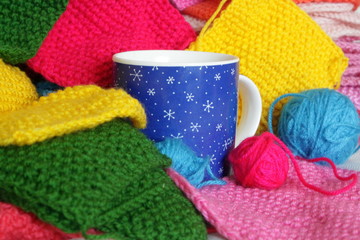 A cup with a drink standing among multicolored  knitted patches and balls of yarn