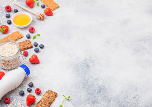 Organic cereal granola bar with berries with honey spoon and jar of oats and bottle of milk drink on stone table background. Space for text.Strawberry, raspberry and blueberry with almond nuts.