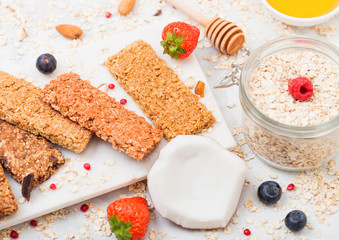 Organic cereal granola bar with berries on marble board with honey spoon and jar of oats and coconut on marble background.