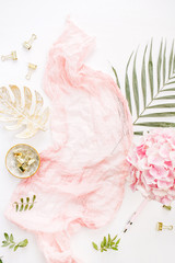 Stylish composition with pink hydrangea flowers bouquet, tropical palm leaf, pastel blanket, monstera leaf plate and accessories on white background. Flat lay, top view.