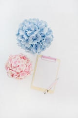 Clipboard mockup and pale pastel hydrangea flowers bouquets on white background. Flat lay, top view minimal home office desk.