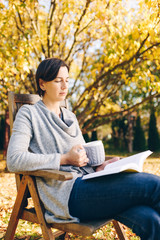 Fototapeta premium Woman wearing warm knit clothes drinking a cup of hot tea or coffee and reading a book outdoors in sunlight - season, literature, leisure time and people concept - beautiful morning in the garden