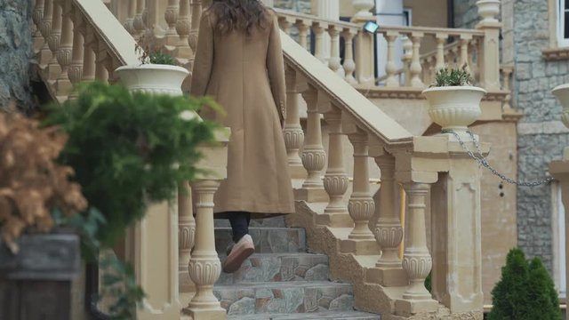 Shooting from the bottom, close-up, slow angle, a young girl dressed in a long cashmere coat, wearing a stylish black hat climbs the steps in a country house.