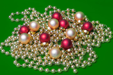 Six small matte red-coloured and six matte golden-coloured Christmas ornaments laying on shiny golden-coloured Christmas beads wreath on green background in studio light