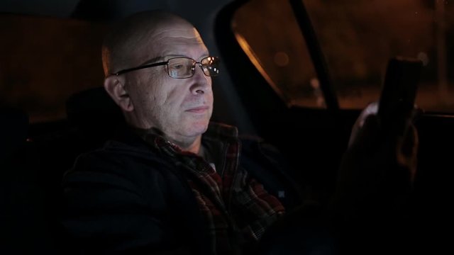 An elderly man using cell phone in a taxi. He is texting, checking mails, chats or the news online. Night.