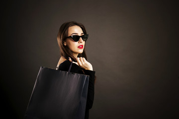 Black friday sale concept. Attractive young woman with long brunette hair, smiling, wearing sexy dress & cat eye sunglasses, holding blank shopping bags over black background. Copy space, close up.