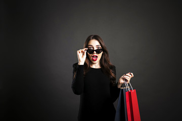 Black friday sale concept. Attractive young woman with long brunette hair, smiling, wearing sexy dress & cat eye sunglasses, holding blank shopping bags over black background. Copy space, close up.