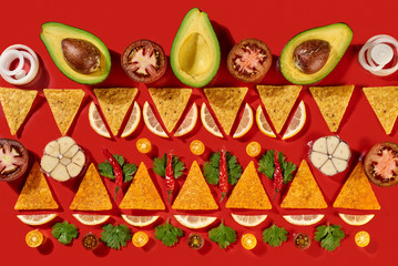 Creative geometric food pattern from mexican nachos corn chips, fresh vegetables, fruits, greens, chili, garlic - ingredients for tomato chili sauce on a red background. Flat lay
