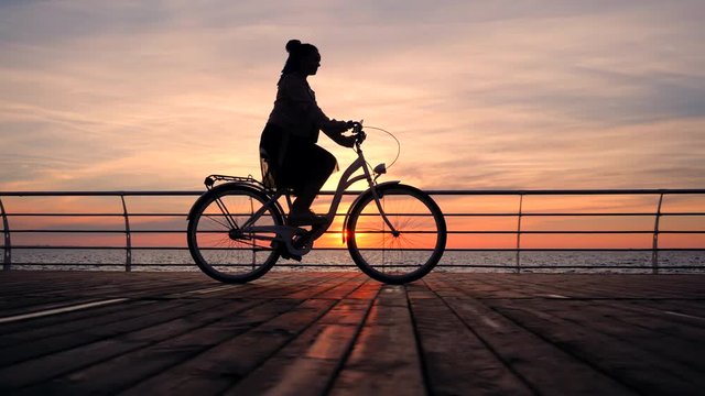 Wonderful sunrise or sunset above ocean. Silhouette of young stylish girl cycling on vintage bike on wooden embankment. Woman on bicycle near sea. Slow motion. 4k.