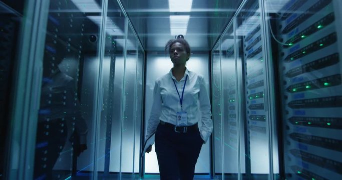 Black woman in formal outfit walking in corridor among glowing server racks in data center