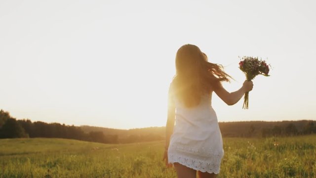Beautiful young woman with wild flower bouquet in white dress running through countryside field at sunset, enjoying freedom in a meadow in sun rays light, happy girl with long less hair run on a field
