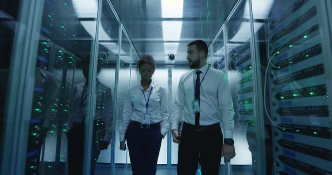 Adult multiracial man and woman with tablets walking in corridor of data center checking server hardware