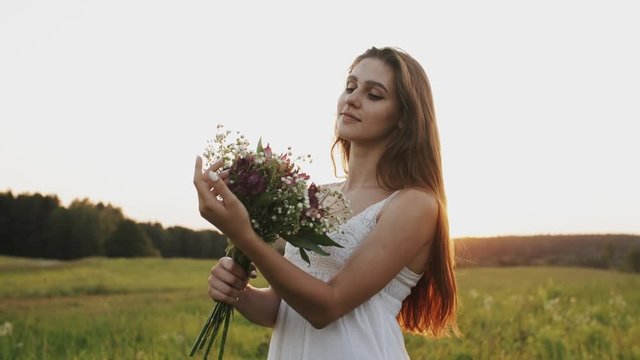 Happy young woman in white dress standing in the field with bouquet of wild flowers with loose long hair in sun rays light at sunset on a summer evening, looking at camera and smiling