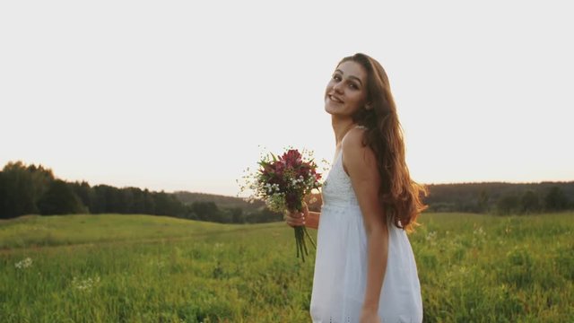 Happy young woman with wild flower bouquet in white dress running through the field at sunset, enjoying and laughing in a meadow in sun rays light, happy girl with long less hair run on a field