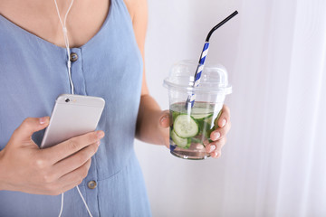 Woman holding plastic cup with tasty fresh cucumber water and mobile phone, closeup