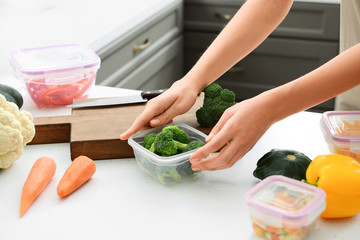 Woman holding plastic container with fresh broccoli for freezing at table in kitchen